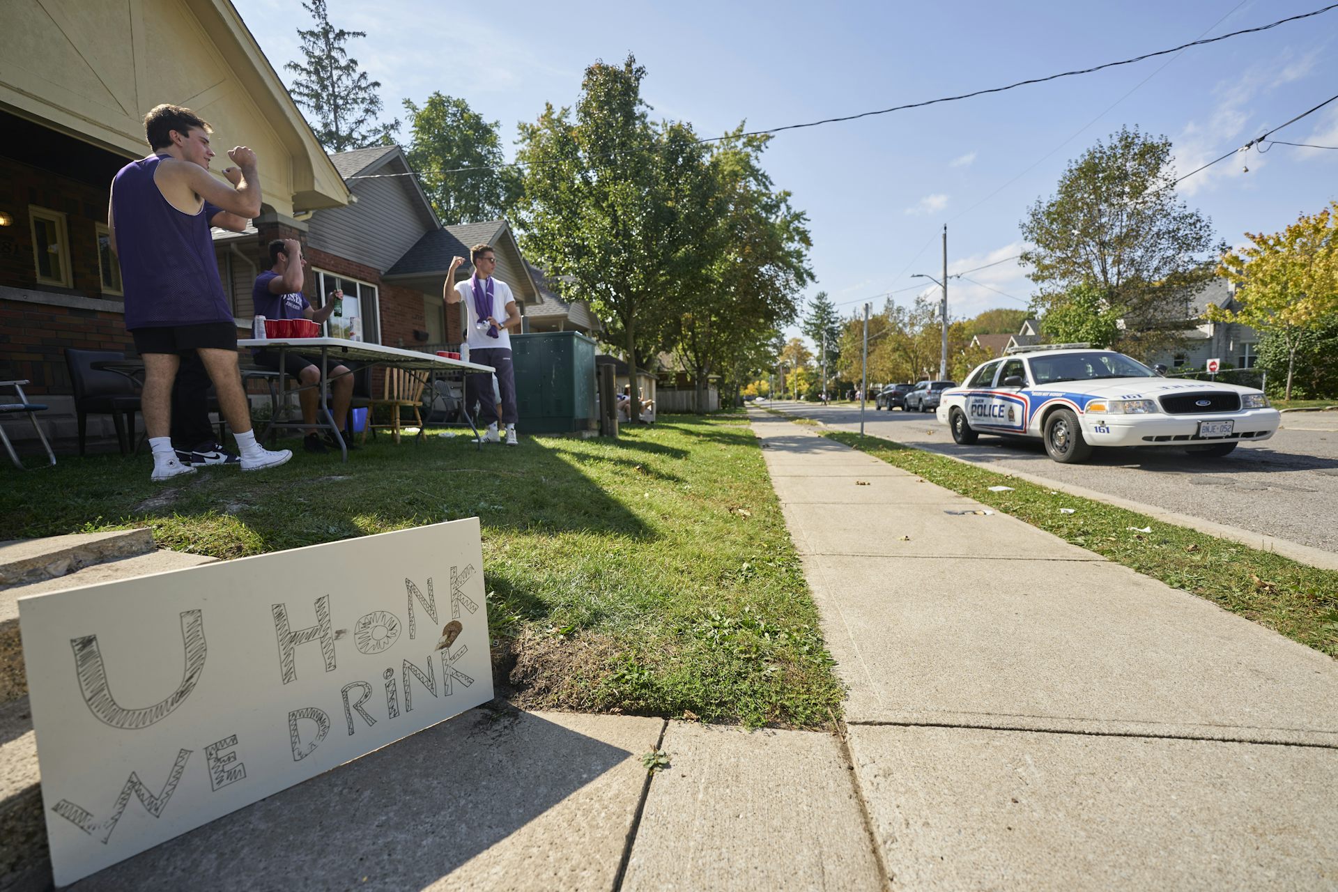 A police car passes a student house where people are drinking outside.