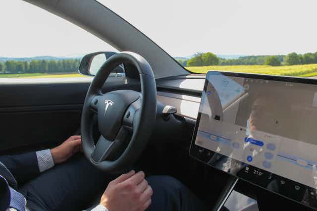Interior of a car showing driver' is hands off the steering wheel, a computer tablet -like display mounted on the dashboard and a natural landscape in the background