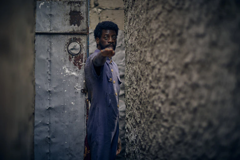 a man with natural hair and a beard and wearing a navy blue overall stands in a doorway of an old building and points a hand directly at camera, looking into the camera.