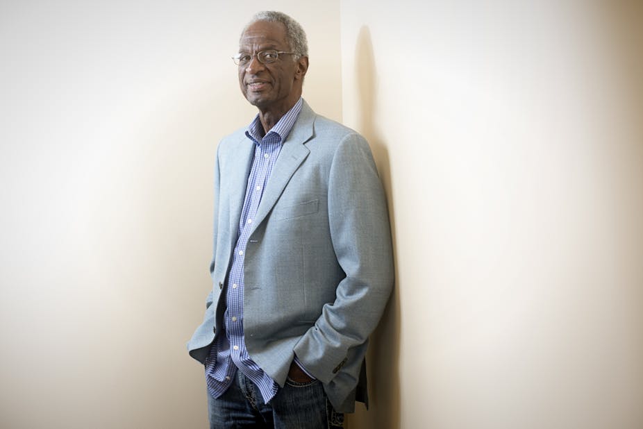 A photo of an older Black man standing in a corner in dress clothes.