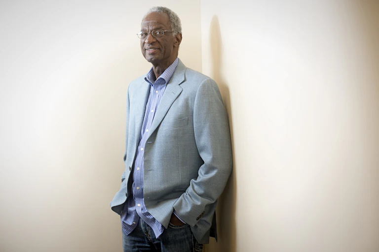 A photo of an older Black man standing in a corner in dress clothes.