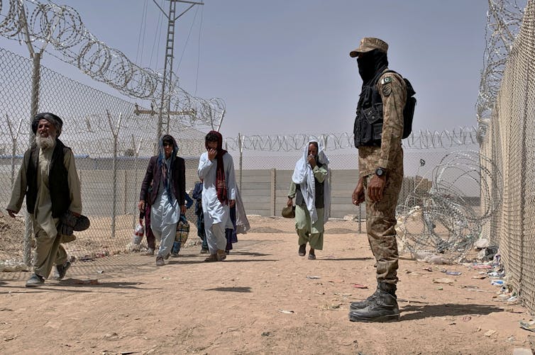 Four Afghan refugees enter Pakistan at a border crossing point marked by barbed wire while a man in army dress watches.
