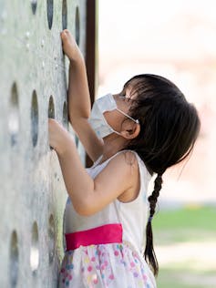 Little girl climbing playground wall with mask on.