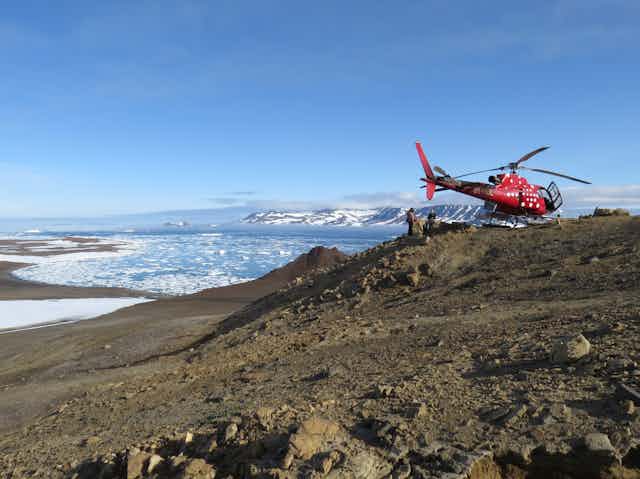 A helicopter parked on a hill with geologists. In the background a small island and packice fills the bay
