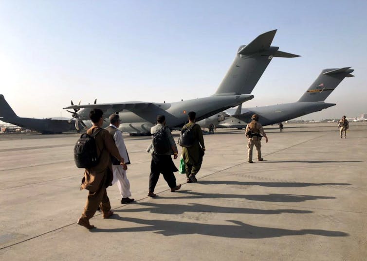 Cinco homens caminhando em direção a aeronaves militares estacionadas em uma pista.