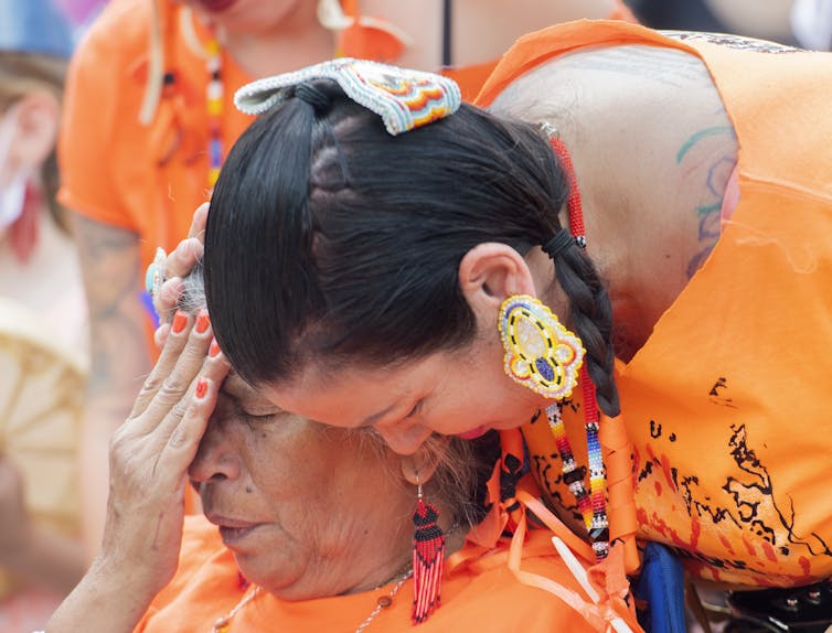 A woman weeps while another consoles her, both wearing orange T-shirts.