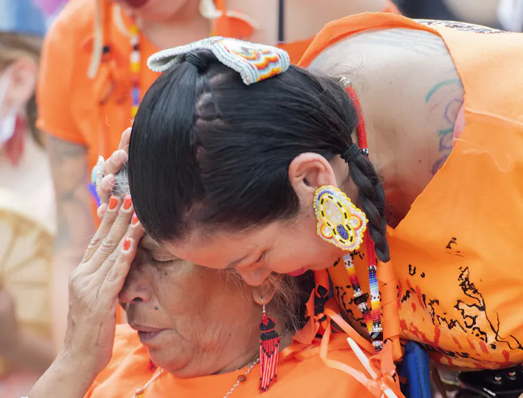 A woman weeps while another consoles her, both wearing orange T-shirts.
