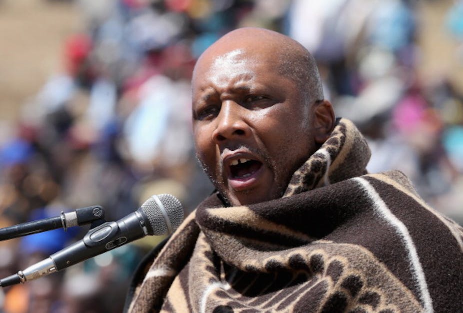 A man wearing a traditional basotho blanketspeaks into two microphones at a public meeting in Lesotho.