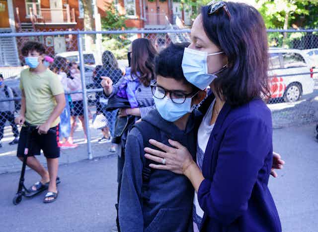A mother embraces her son in a school yard, both wearing face masks.