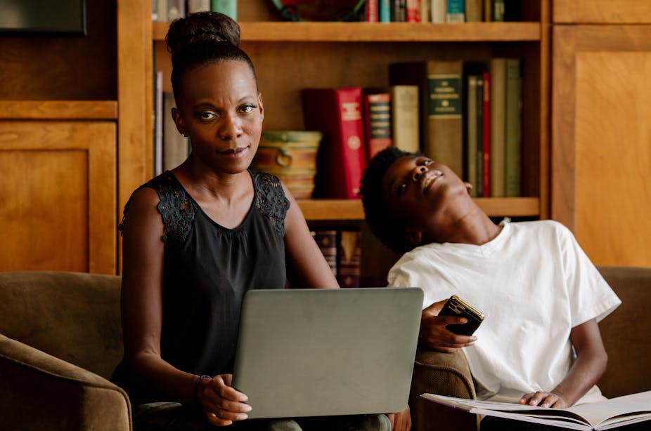 A Black mother holding a laptop computer in her living room looks exasperated while her tired-looking preteen son sits nearby.