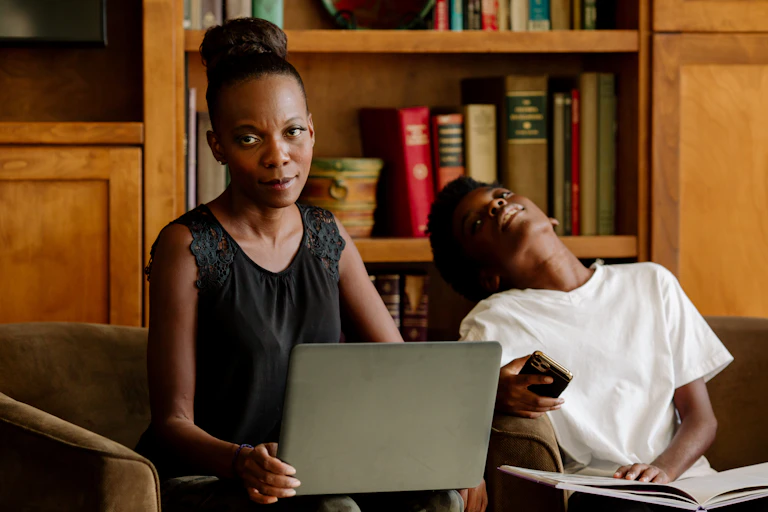 A Black mother holding a laptop computer in her living room looks exasperated while her tired-looking preteen son sits nearby.