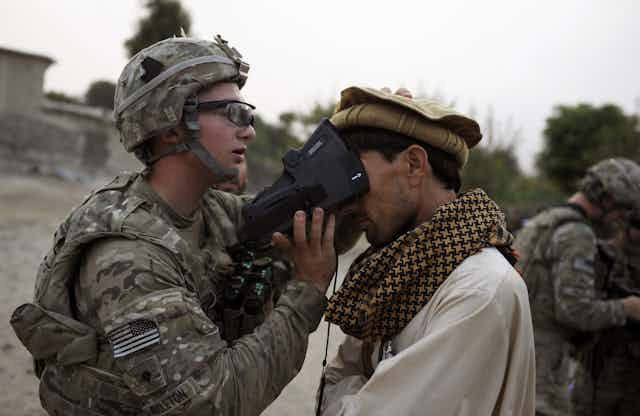 A soldier in battle armor with a United States flag shoulder patch presses a device against the upper face of a man wearing a large scarf and a traditional Afghan hat