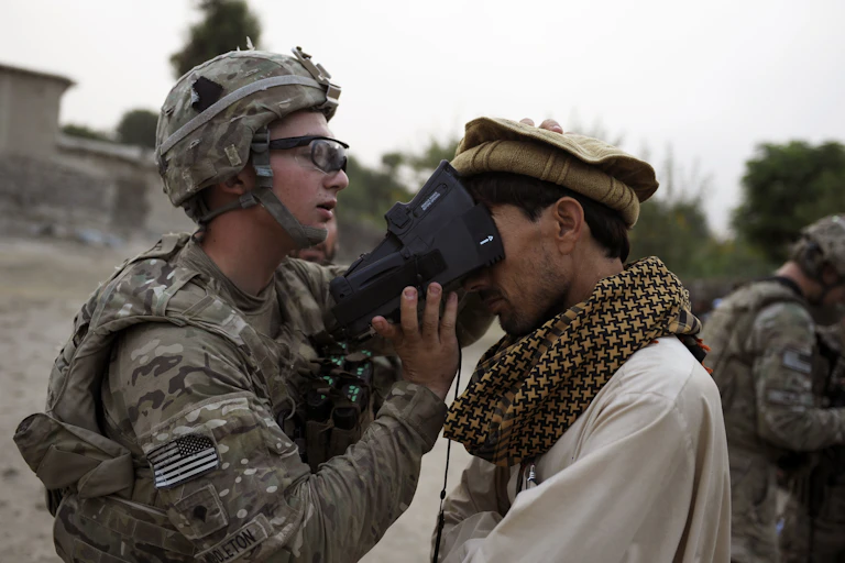 A soldier in battle armor with a United States flag shoulder patch presses a device against the upper face of a man wearing a large scarf and a traditional Afghan hat.