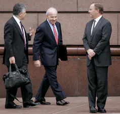 Two men – one carrying a bag, another a portfolio – are walking and appear to be talking toward another man who is standing and looking at them with folded hands.