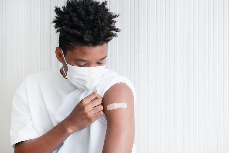 A teenage boy with a mask on and a band-aid on his arm.