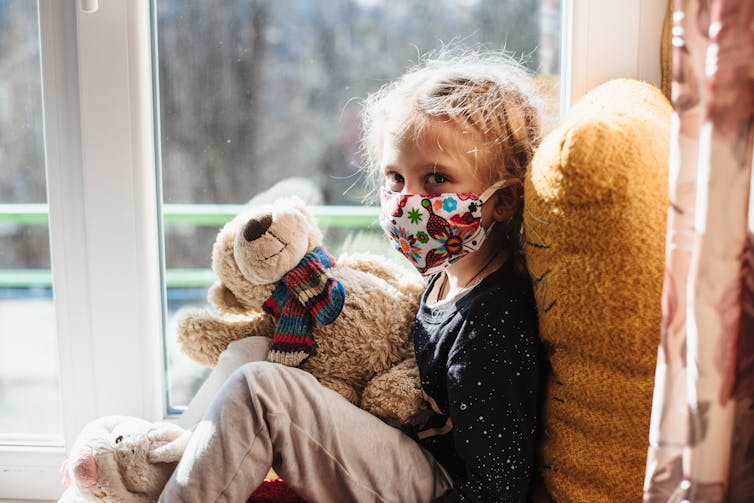 A young girl in a mask with a teddy bear.