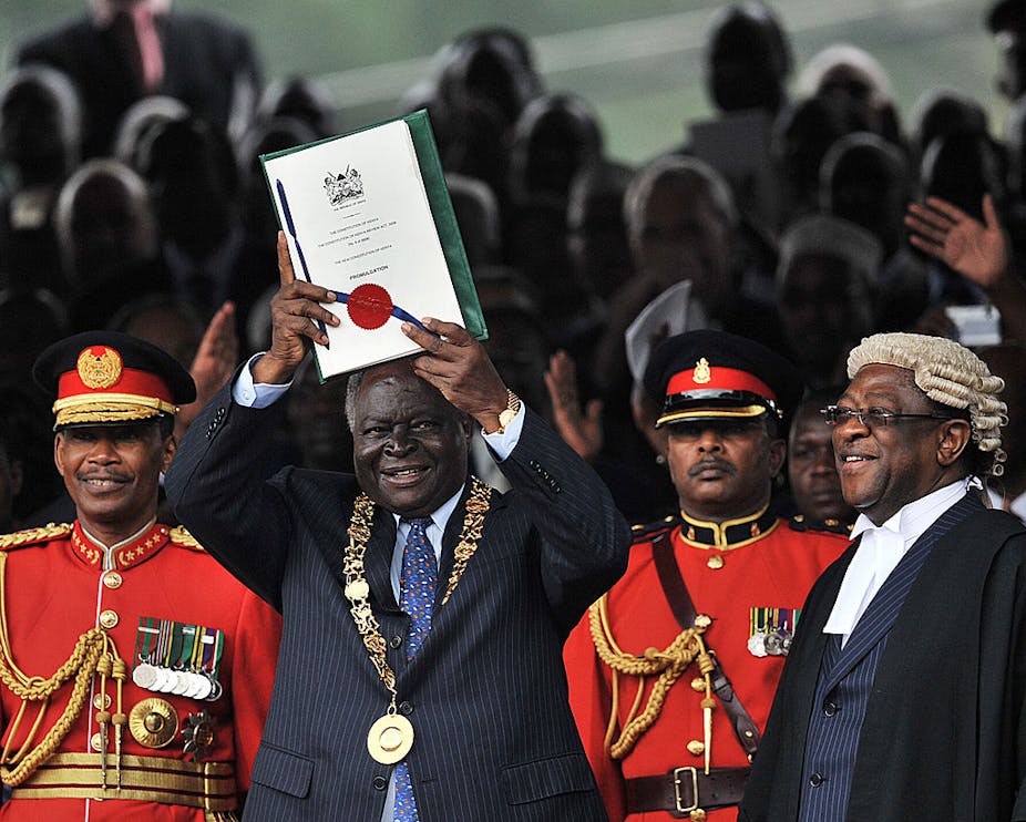 Man wearing a ceremonial gold chain holds up a document with a seal and ribbon, while a man in a robe and white wig and men in military uniform stand nearby