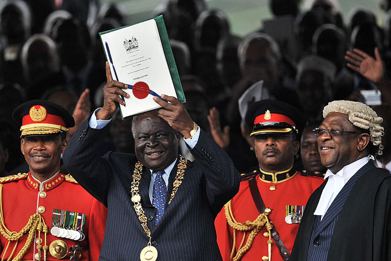 Man wearing a ceremonial gold chain holds up a document with a seal and ribbon, while a man in a robe and white wig and men in military uniform stand nearby