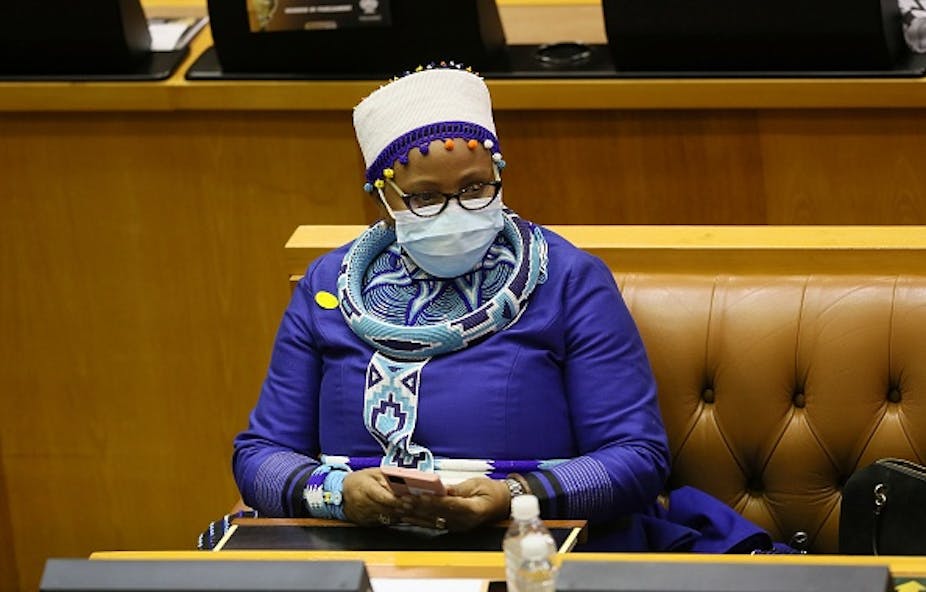 A woman wearning a hat and traditional Xhosa attire holds a cellphone in both hands while sitting on a leather bench in Parliament.