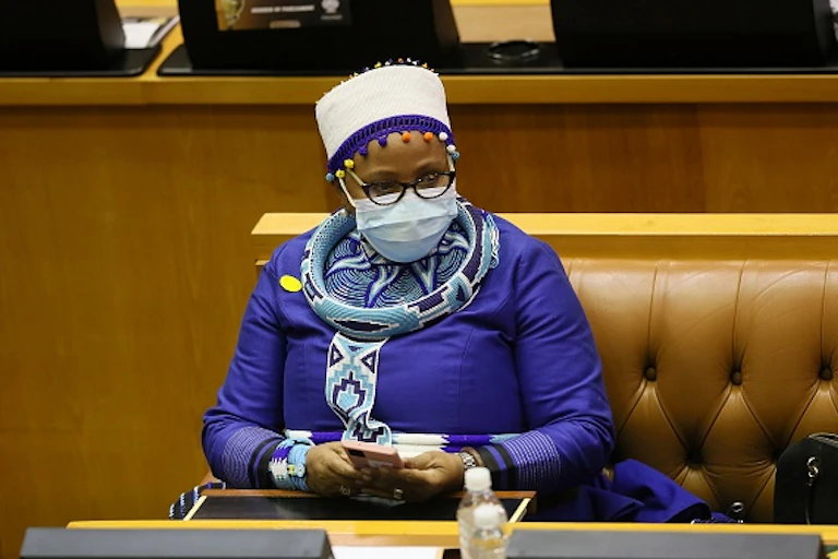A woman wearning a hat and traditional Xhosa attire holds a cellphone in both hands while sitting on a leather bench in Parliament.