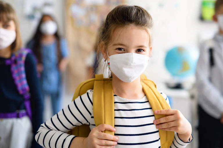 A young girl at school, wearing a backpack and a facemask