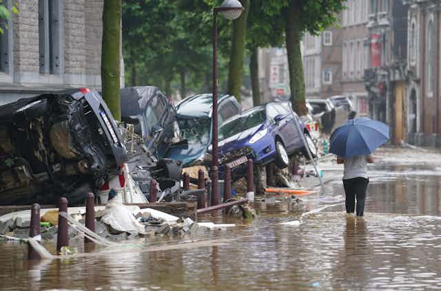 A person with an umbrella walks knee-deep in water down a flooded street with cars overturned and piled up on the sides.