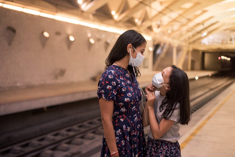 Mother and daughter listening to music and wearing a mask while waiting for train.
