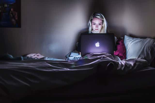 Young woman focuses intently on laptop in bed, her face lit up by the screen