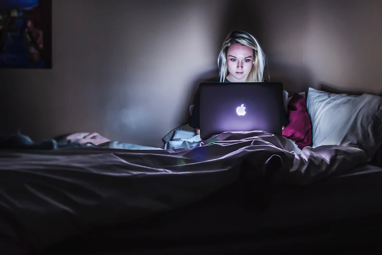Young woman focuses intently on laptop in bed, her face lit up by the screen