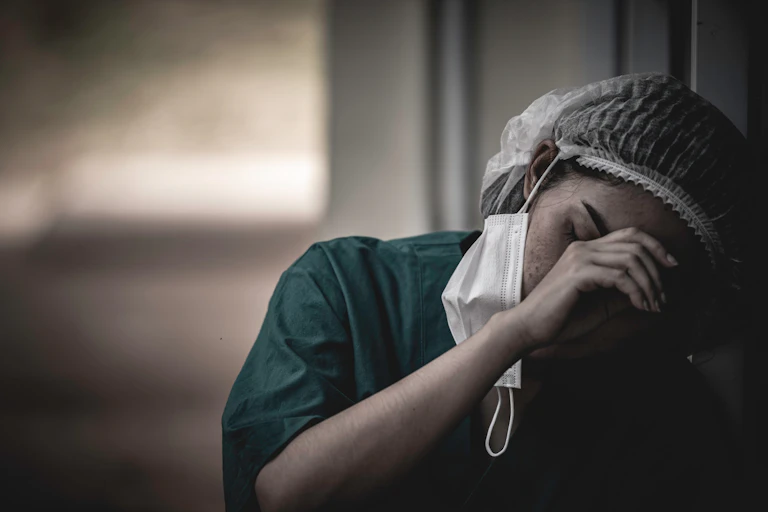 Exhausted nurse with face mask loosened slumped against a wall looking exhausted.