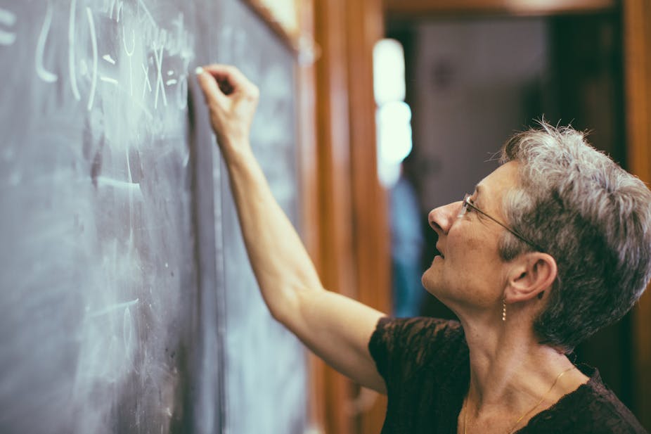 Maths teacher with grey hair writing on a chalkboard.