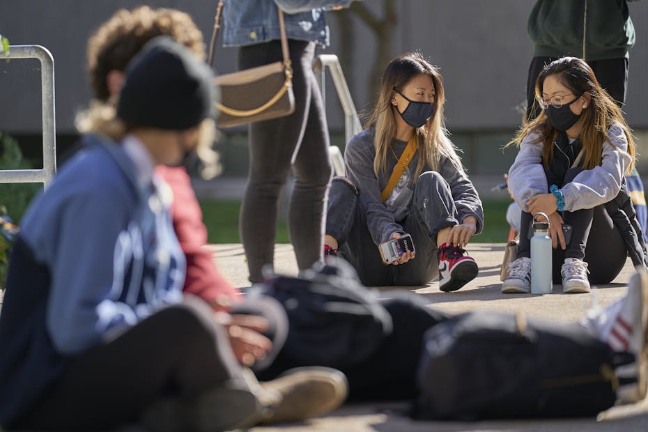 Masked people sit on the pavement in the sun.