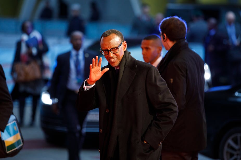 A bespectacled man dressed in a coat raises his hand in greeting while standing amid other people