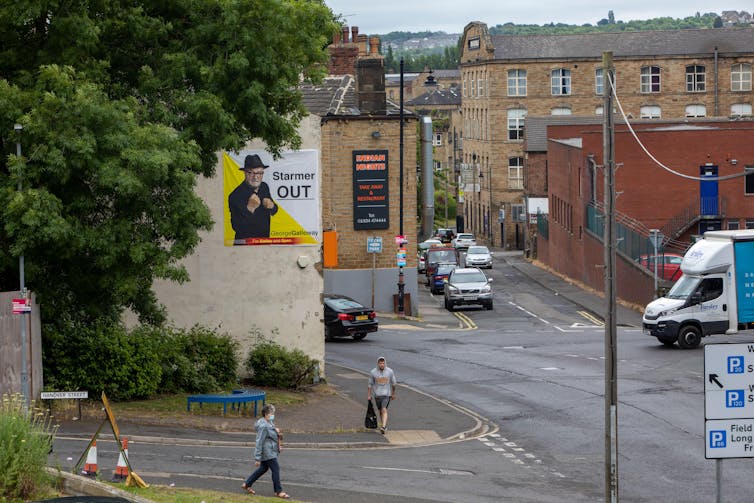 A view of a street in Batley and Spen with a large poster of George Galloway hanging on the side of the building. Galloway has his fists up in the picture, next to some text reading 'Starmer out'.