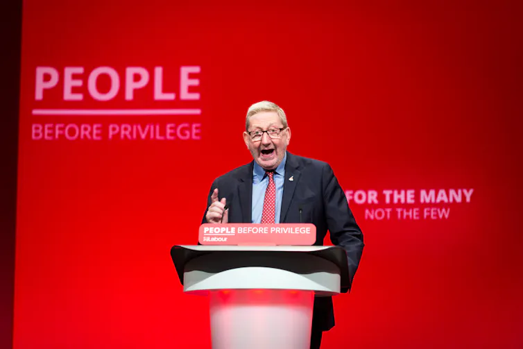 Len McCluskey delivering a speech in front of a sign reading 'people over privilege'.