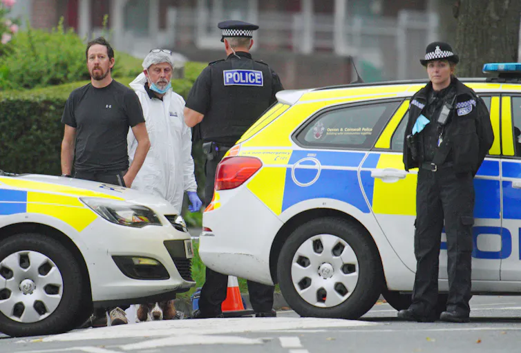 Police officers and a man in forensic gear stand in front of vehicles blocking off a suburban street.