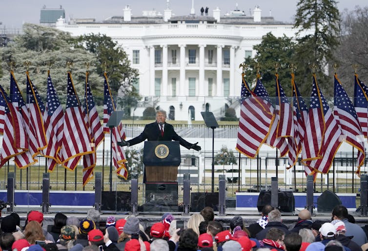 A man in a suit stands with his arms spread on a platform lined with U.S. flags, in front of the White House and a crowd of people