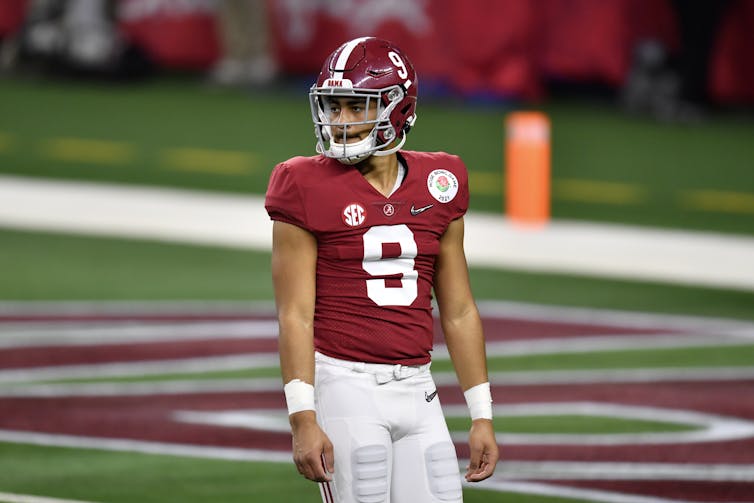 A man wearing a red football jersey with the number 9 on it stands on a football field facing the viewer's left.