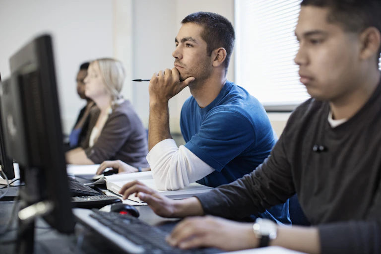 Four young people sit at a long table with computer keyboards and monitors in front of them