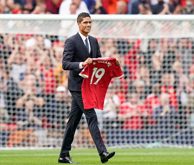 Varane holding his new Manchester United shirt on the pitch.