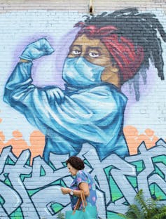 A woman walks past a street mural of a health care worker in protective gear flexing his biceps