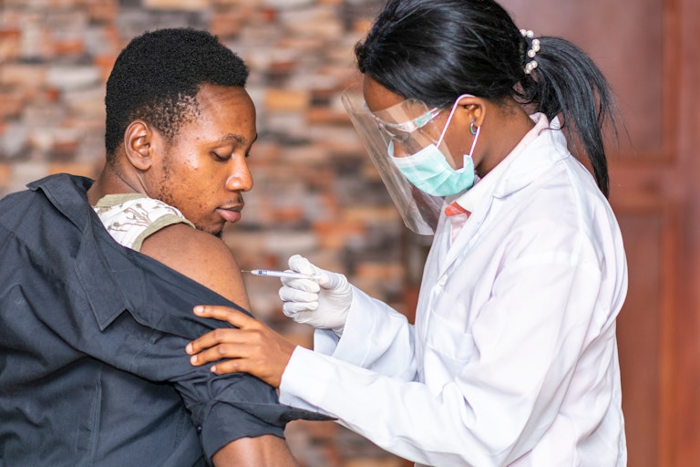 A woman in a white coat, wearing a surgical mask and a face shield, is giving a man an injection in his shoulder. He is watching the needle, looking over his shoulder, and appears interested.