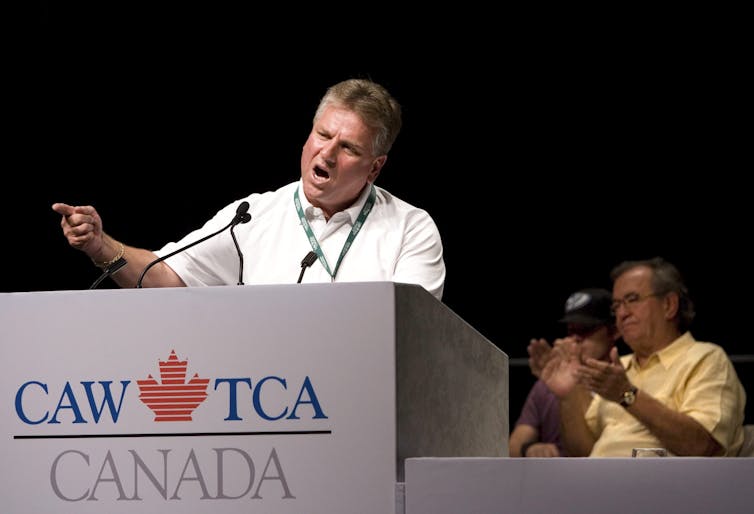 Ken Lewenza gestures while standing behind a dias that has the CAW logo.