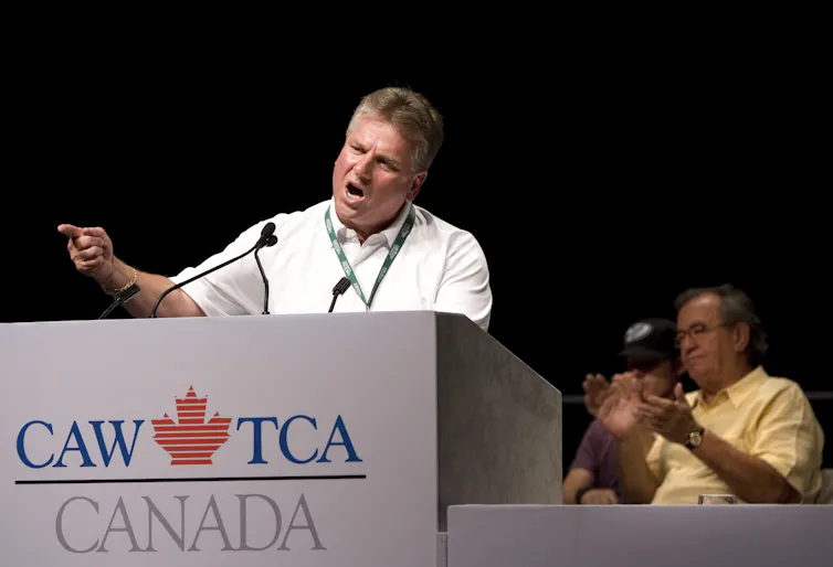 Ken Lewenza gestures while standing behind a dias that has the CAW logo.