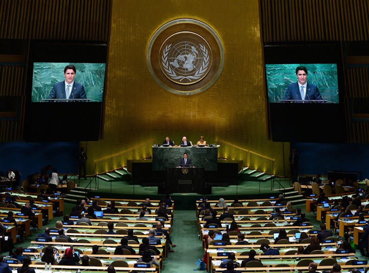 Trudeau delivers a speech and is seen on jumbo screens to the left and right of him in a ornate hall.
