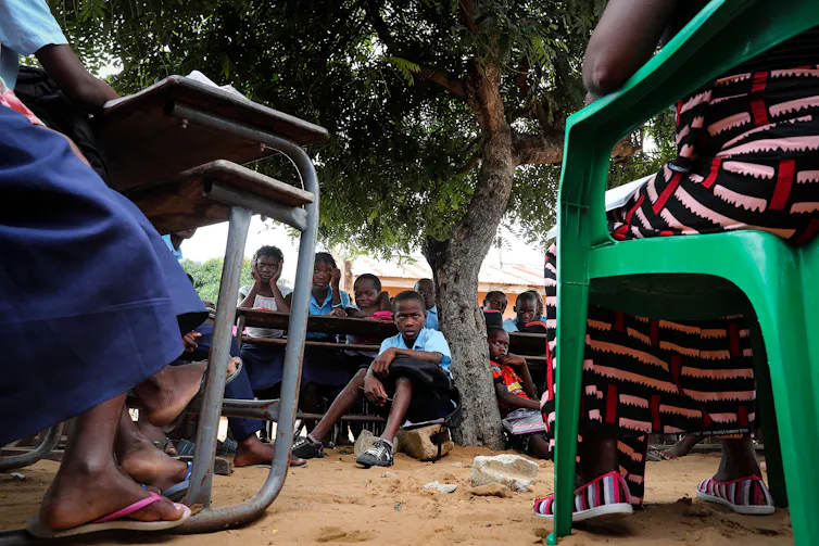 Children attend a class sitting on the ground under a tree.