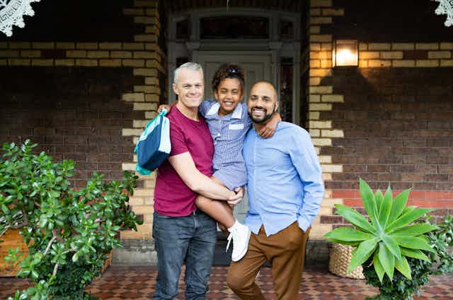 Two dads hold school-aged daughter while standing in front of house 