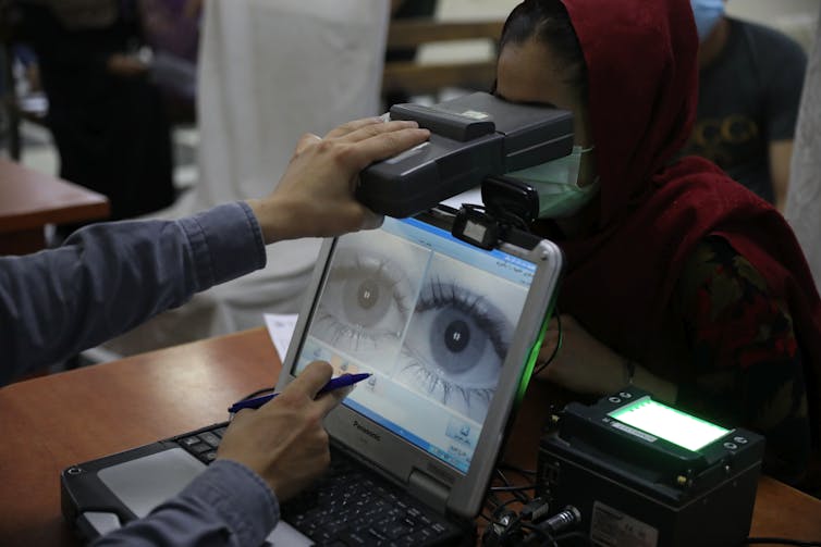 La pantalla de una computadora muestra una imagen ampliada de un par de ojos mientras un brazo sostiene un objeto en forma de caja frente a los ojos de una mujer que usa un pañuelo en la cabeza y una mascarilla.