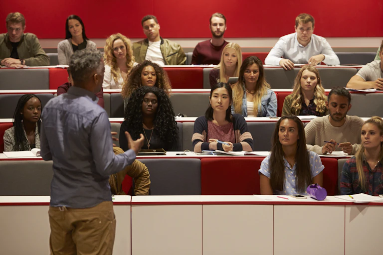 A man stands with his back to the camera giving a lecture to people sitting in front of him.