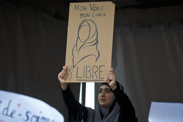 A woman in a headscarf at a protest in Toulouse, France, shows a drawing reading 'My veil, my choice, Free' in French.